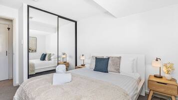 A modern hotel room with a large bed, a bedside table with a lamp, a mirror, and a wardrobe.