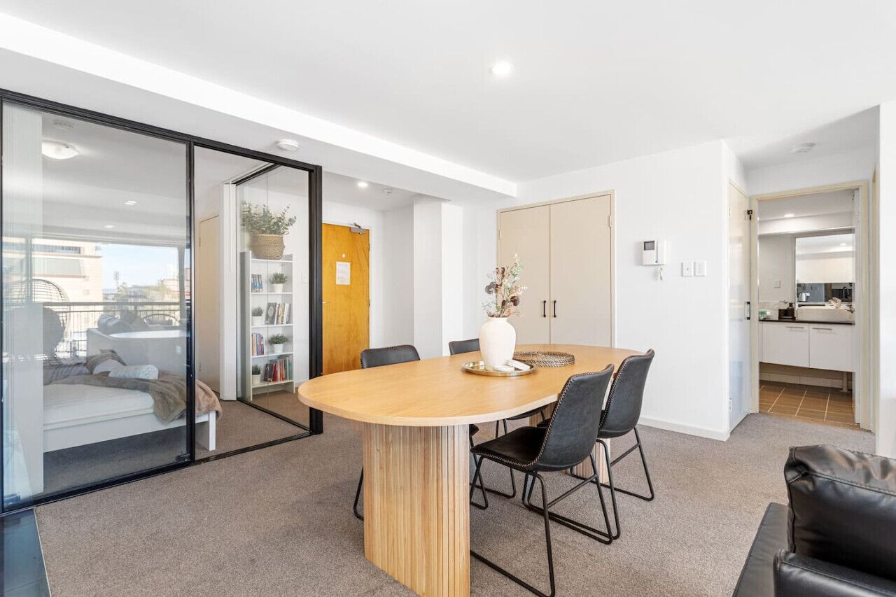 A modern living room with a wooden dining table, black chairs, a glass partition, a bookshelf, and a view of the city.
