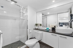 Une salle de bain moderne avec une douche à parois de verre, des toilettes blanches, un lavabo avec un miroir et un plan de toilette où sont posés les articles de toilette.