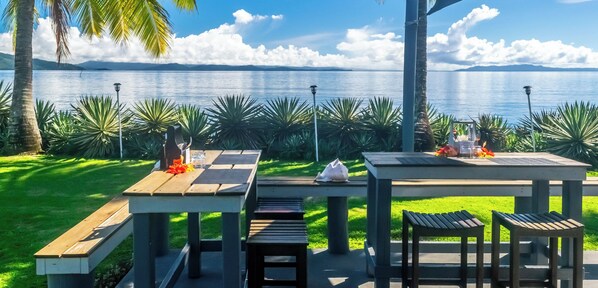 An outdoor dining area with a table and benches, surrounded by tropical plants and a view of a lake.