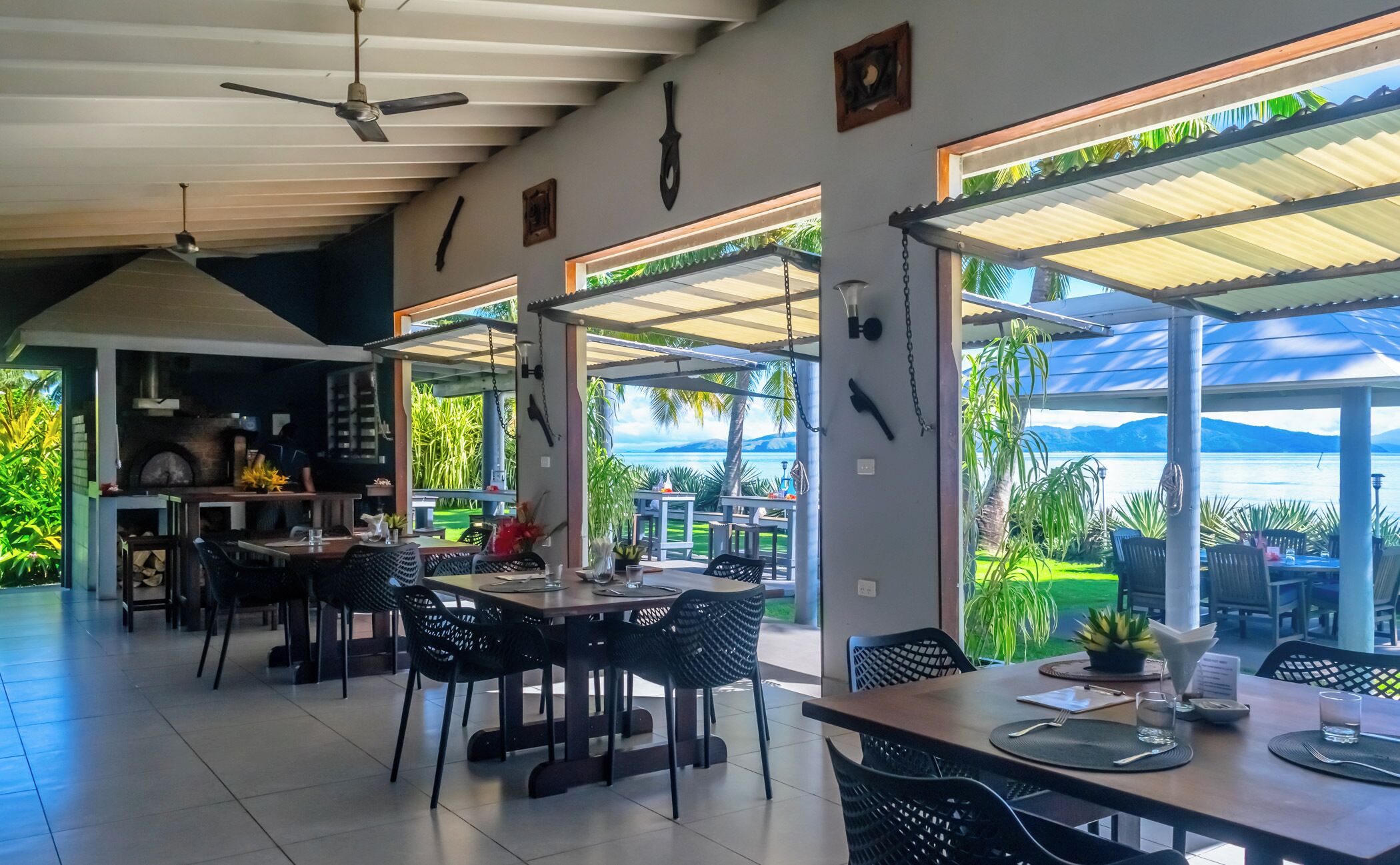 Un vaste espace de salle à manger avec vue sur la mer, doté d’un haut plafond, de ventilateurs de plafond et d’un coin cuisine à l’arrière-plan.