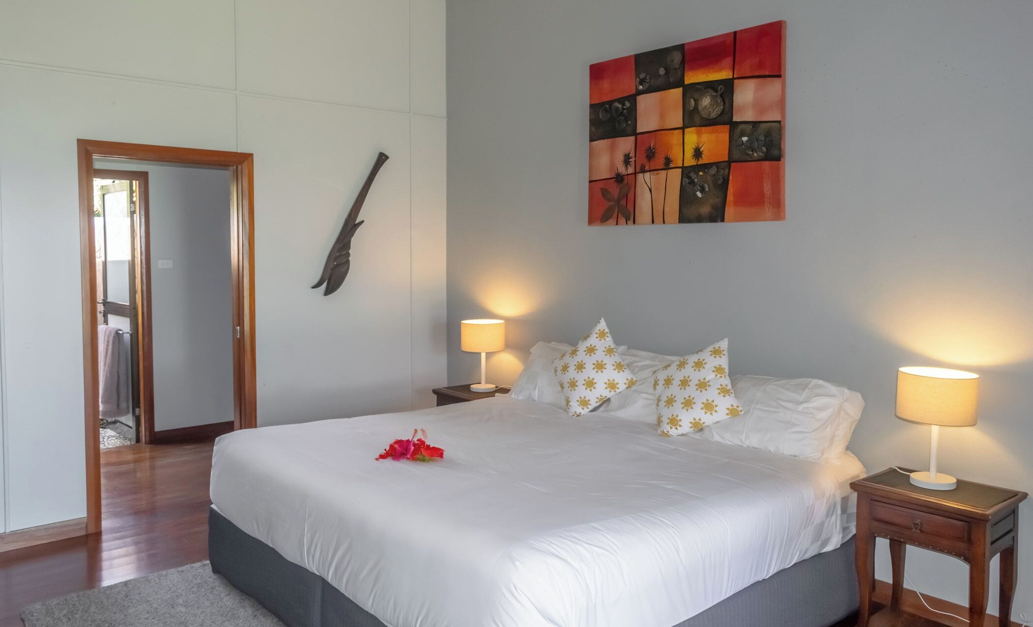 Un lit bien fait, avec du linge de lit blanc et des coussins ornés de motifs solaires, deux lampes de chevet et une peinture abstraite aux couleurs vives accrochée au mur.
