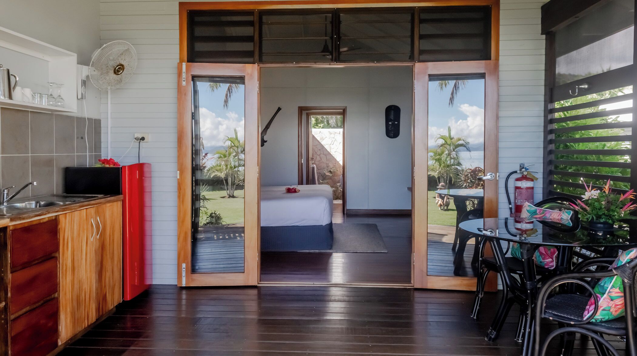 A room with a bed, a small table, and a fan. There is a view of a garden outside.