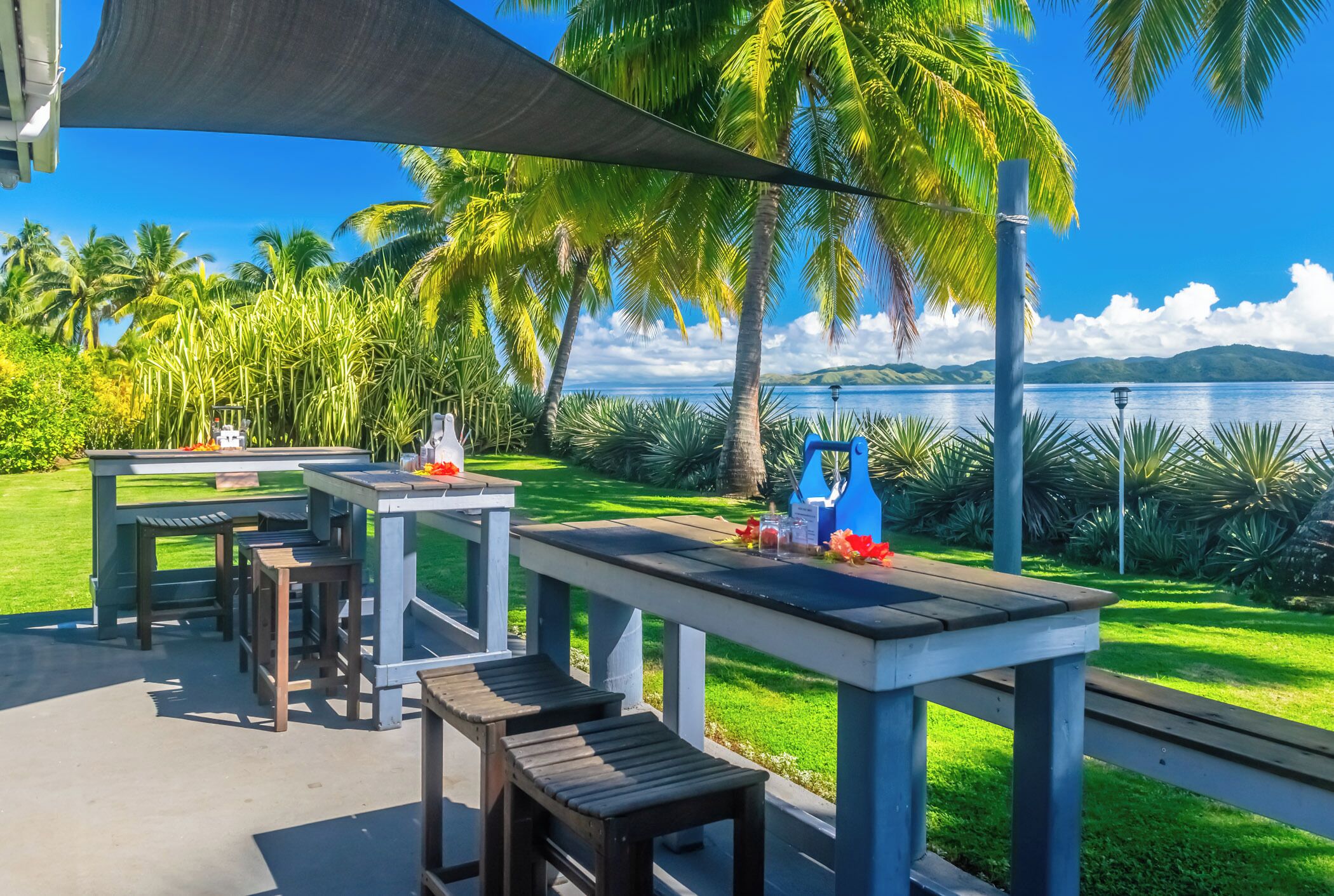 An outdoor patio area with tables and benches, overlooking a body of water and palm trees.