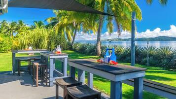 An outdoor patio area with tables and benches, overlooking a body of water and palm trees.