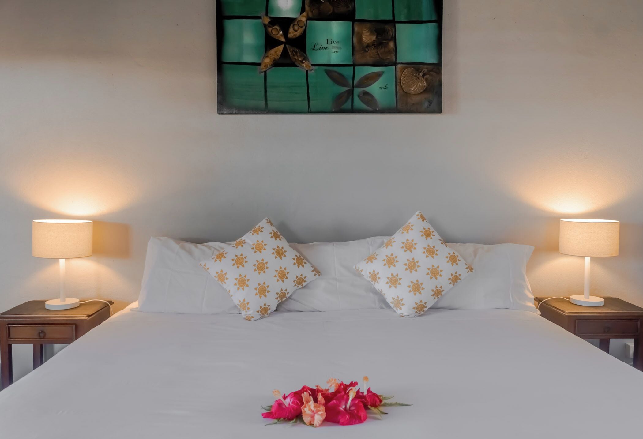 A neatly made bed with white sheets and two pillows, a floral arrangement on the bed, and a wall art piece above.