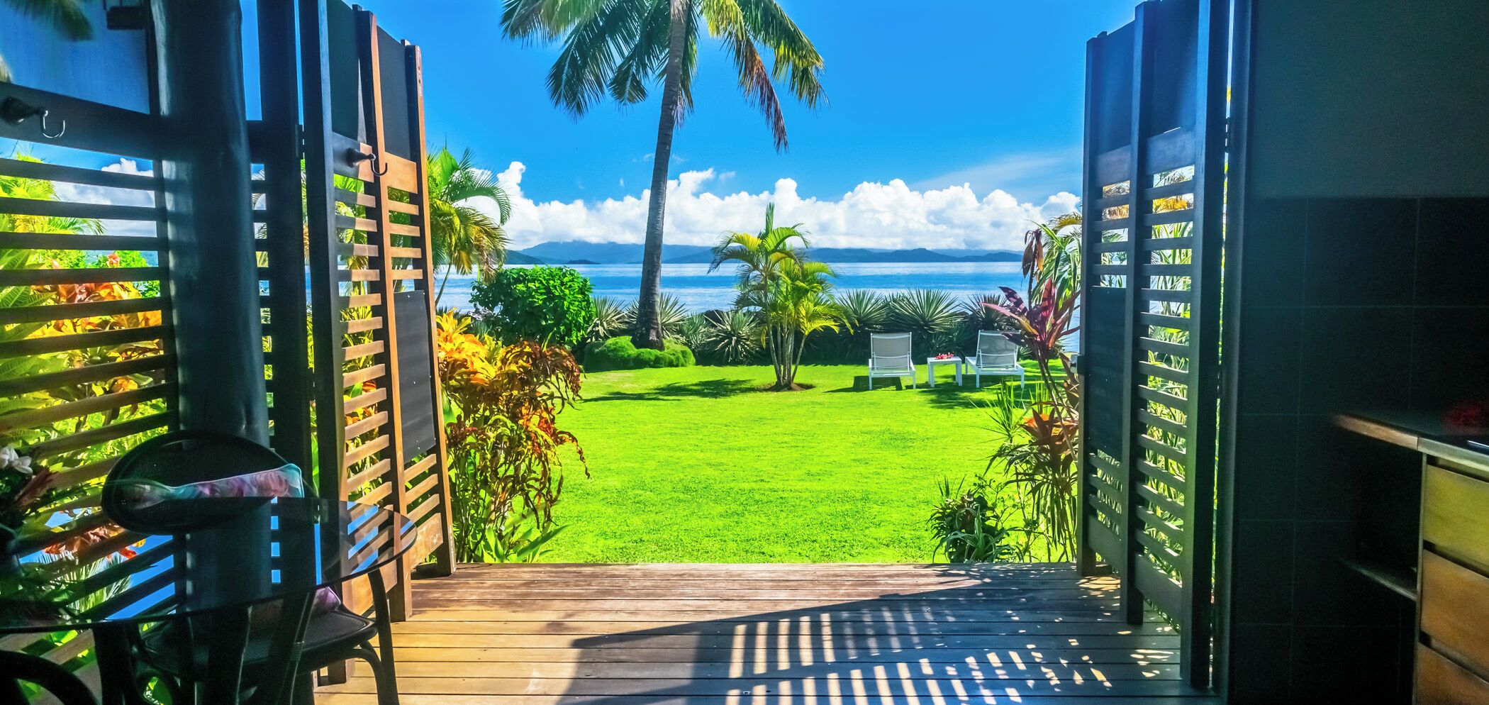 A view from a covered outdoor area to a grassy lawn with a pool and lounge chairs, overlooking a body of water and palm trees.