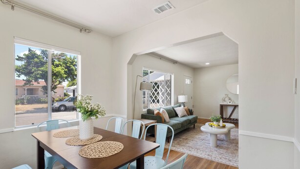A dining area with a wooden table, blue chairs, and a vase with flowers. There is a sofa with cushions and a lamp in the background. - Modern Coastal Retreat – 3BR Gem in Long Beach (Long Beach)
