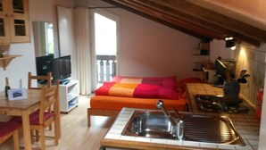 A compact living space with a kitchenette, a sofa with colorful cushions, a dining table with chairs, a television, and a small balcony.