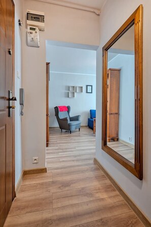 A hallway with wooden flooring, a large mirror on the right, and a chair with a red cushion in the background.