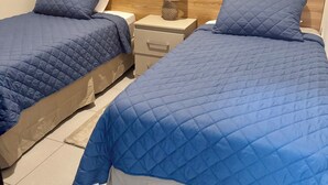A hotel room with two beds, each with a blue quilted bedspread and matching pillows. There is a wooden headboard, a bedside table with a lamp, and a nightstand. The room has a tiled floor and artwork on the walls. - Beautiful house in the most exclusive área of San Miguel (San Miguel)