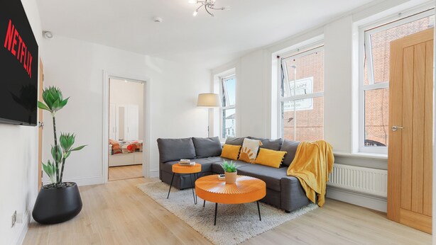 Ein modernes Wohnzimmer mit einem grauen Sofa, einem kleinen orangefarbenen Tisch, einer Topfpflanze und einem großen Fenster mit Blick auf ein Backsteingebäude.