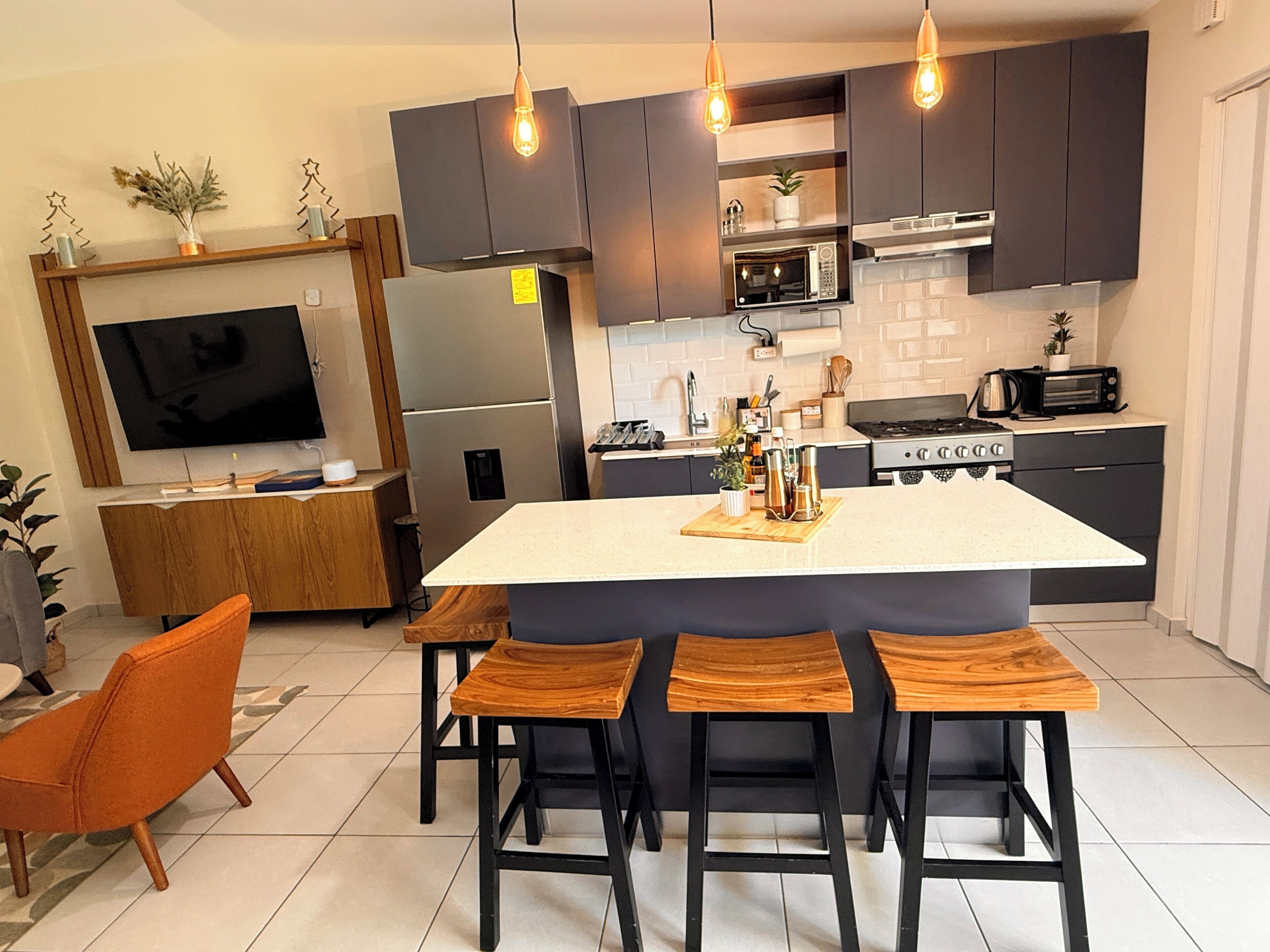 Una cocina moderna con una isla central, taburetes de bar, horno empotrado, microondas y un televisor montado en la pared.