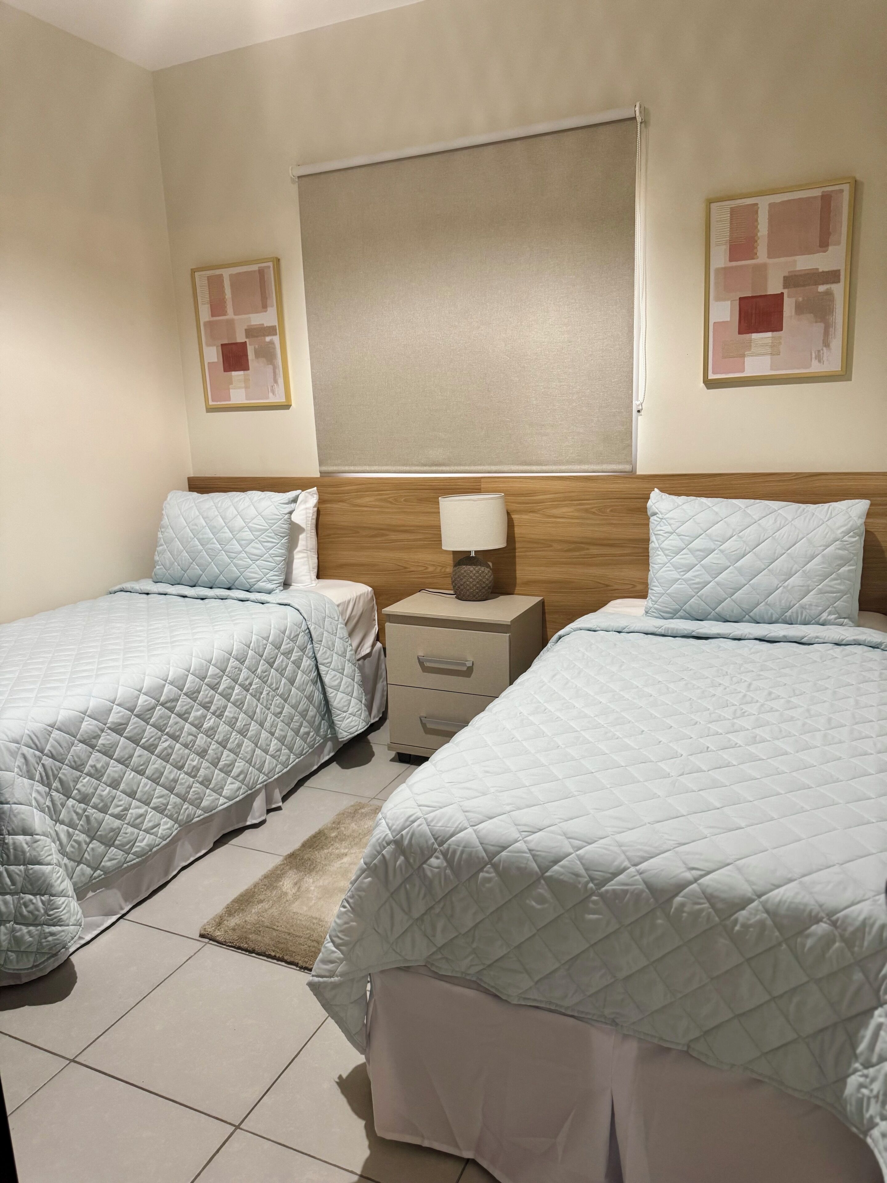 Een hotelkamer met twee bedden, elk voorzien van een gewatteerde dekbedovertrek en kussens, een nachtkastje met een lamp en een groot raam met een blind.