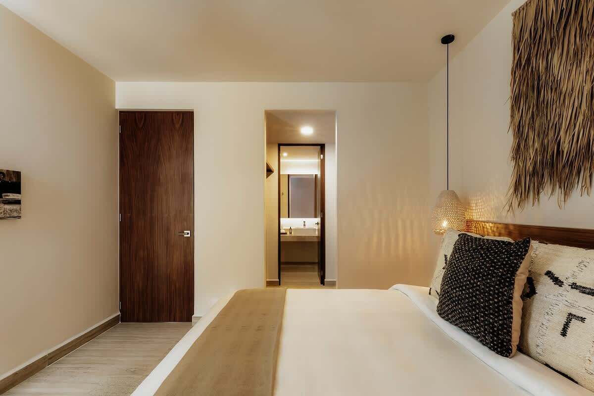 A hotel room with a wooden door, a bed with white bedding, a hanging light fixture, and a visible bathroom in the background.
