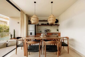 A dining area with a wooden table and chairs, a hanging light fixture, and a kitchen in the background. - Baya Samsara Jungle Paradise Beach Club Access (Tulum)