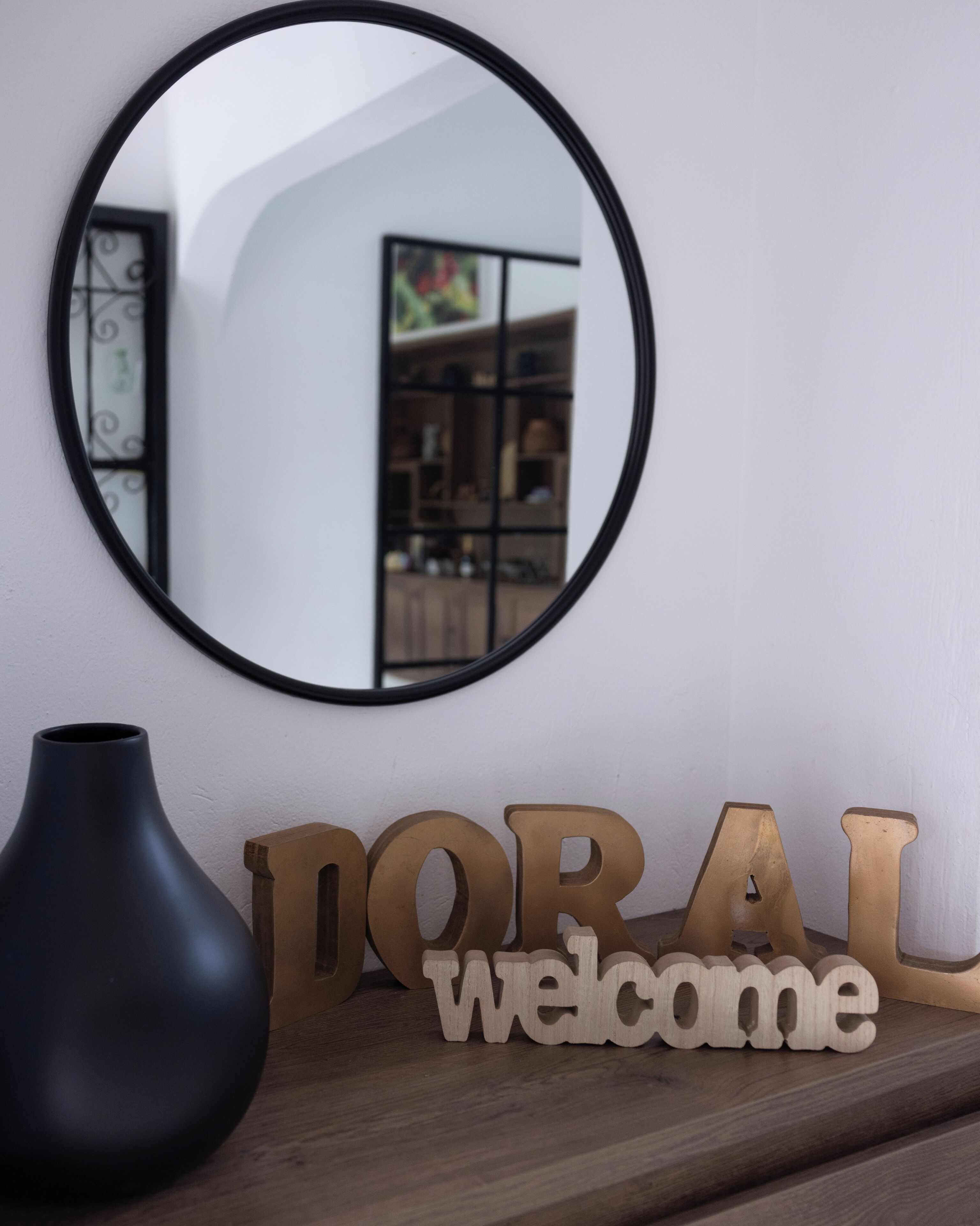 A round mirror on a wall, a dark vase, and wooden letters spelling "DORAL welcome" on a wooden surface.