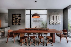 A modern dining area with a wooden table, six stools, a hanging pendant light, and wall art.