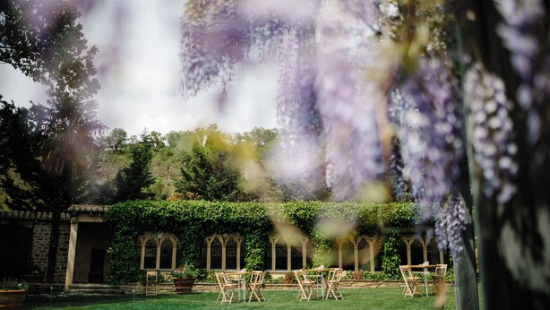 Un jardín al aire libre con un edificio de piedra, ventanas de arco y un área de estar con sillas y mesas.