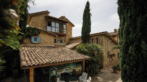 Un edificio de piedra tradicional con techo de tejas, un patio acristalado con mobiliario de mimbre y un ciprés alto.