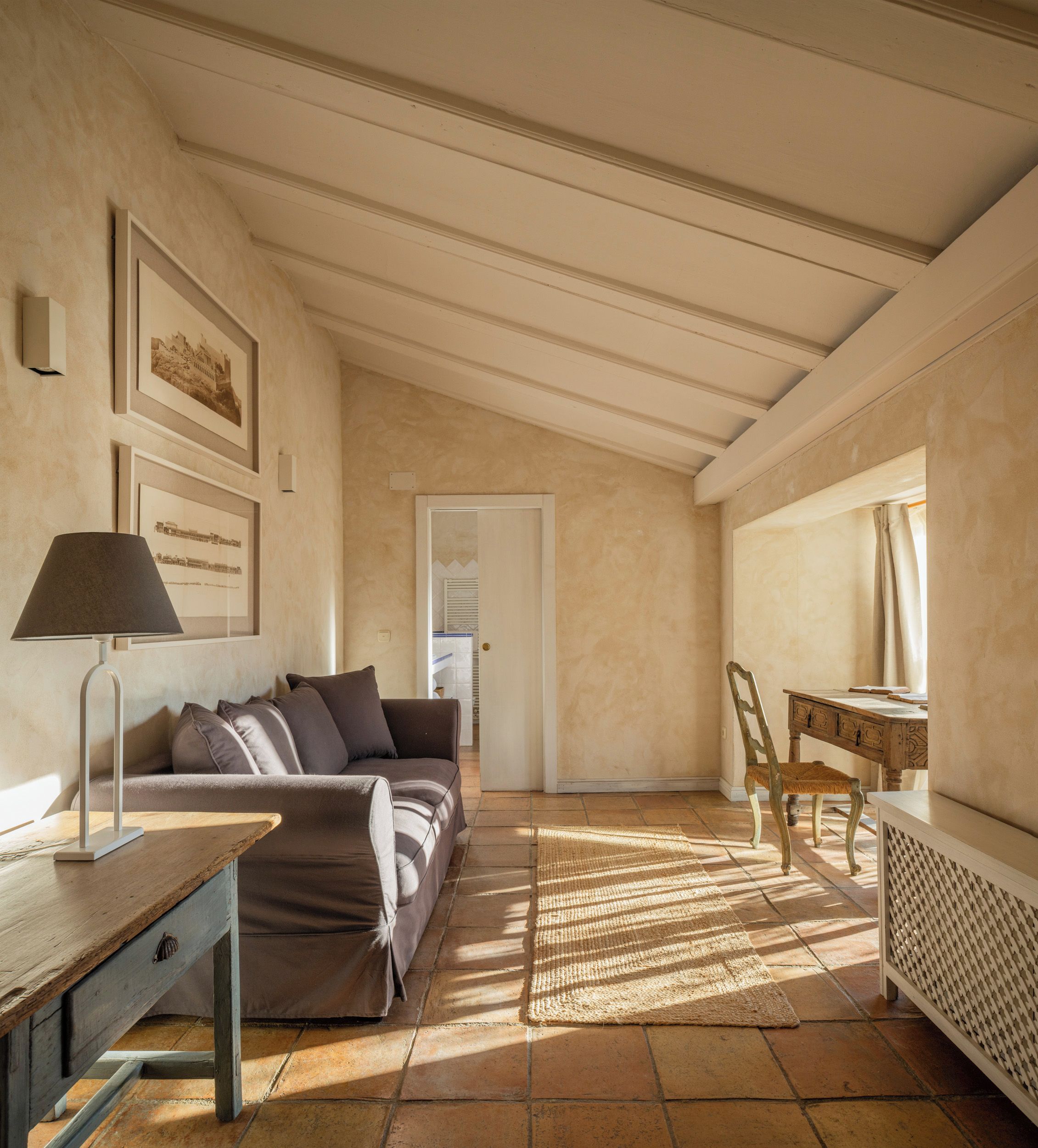 Habitación con sofá, lámpara, silla, mesa y radiador. Se observan cuadros enmarcados en la pared y una puerta que da a otra habitación.