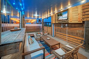 An outdoor patio area with wooden furniture, a surfboard, and a TV mounted on a wooden wall. - Salty Texan by Avantstay Central Location, Views (Port Aransas)