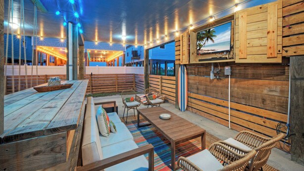 An outdoor patio with wooden furniture, a surfboard, and a TV mounted on a wooden wall. - Salty Texan by Avantstay Central Location, Views (Port Aransas)
