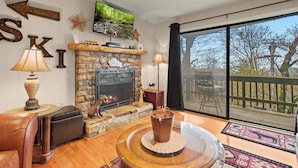 A cozy living room with a stone fireplace, leather sofa, and a large window overlooking a deck. - Tranquility Beech: 2BR Condo Steps from the Resort--Parking Lot Level: No Stairs (Banner Elk)
