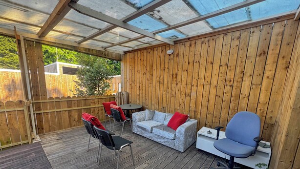 A covered patio with a wooden deck, a grey sofa with red cushions, a small round table, and a blue office chair. - Spacious 4BR Bungalow with Parking Near Spider-Man Filming Locations (London)