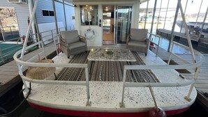 Una terraza techada con asientos y una pequeña mesa, una puerta de vidrio que da al interior, y vista al agua.