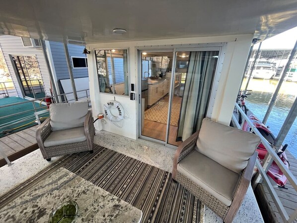 Una terraza cubierta al aire libre con mobiliario de mimbre, una mesa y un ventanal corredero que da a un espacio interior con cocina y zona de estar.