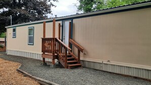 Une maison mobile de plain-pied, avec un escalier en bois menant à une porte, un petit porche et une allée gravillonnée.