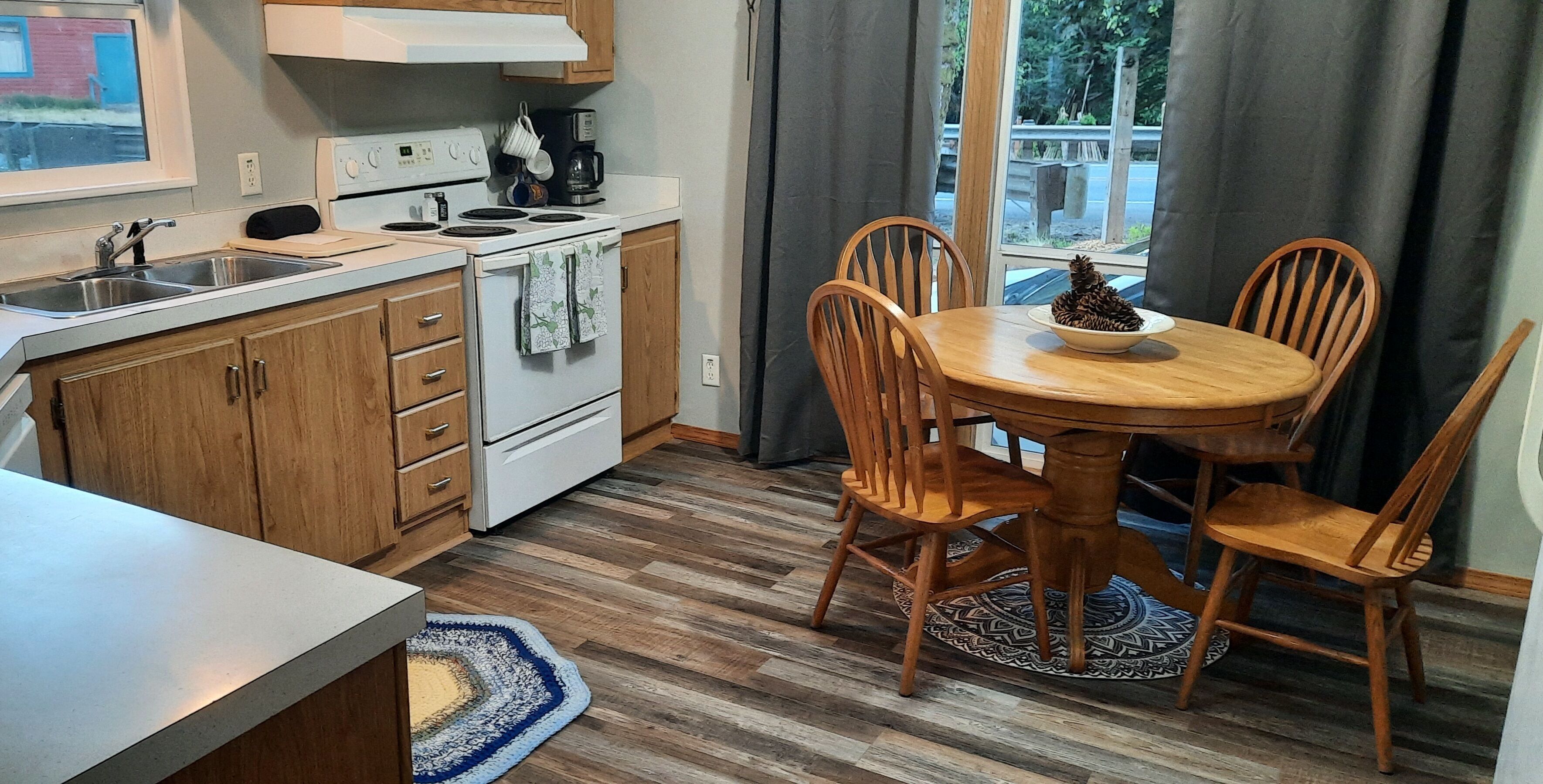 Une cuisine compacte équipée d’une cuisinière, d’un évier et de meubles de rangement en bois. On y trouve une table à manger avec des chaises et une fenêtre agrémentée de rideaux.