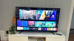 En platt-TV placerad på en vit TV-bänk, med en fjärrkontroll bredvid. På skärmen visas olika streamingtjänster och en blomvas står på bänken.
