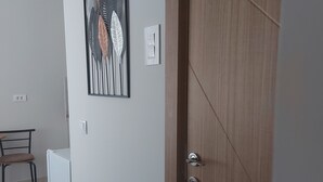 A room with a wooden door, a white wall, a framed artwork, a chair, a small table, and a power outlet. - centrocentro (Novi Sad)