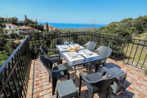 A terrace with a table set for two, surrounded by wicker chairs, overlooking a coastal landscape.