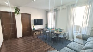 Ein modernes Wohnzimmer mit einem grauen Sofa, einem Essbereich mit Tisch für vier Personen, einem wandmontierten Flachbildfernseher und einem großen Fenster mit Vorhängen.