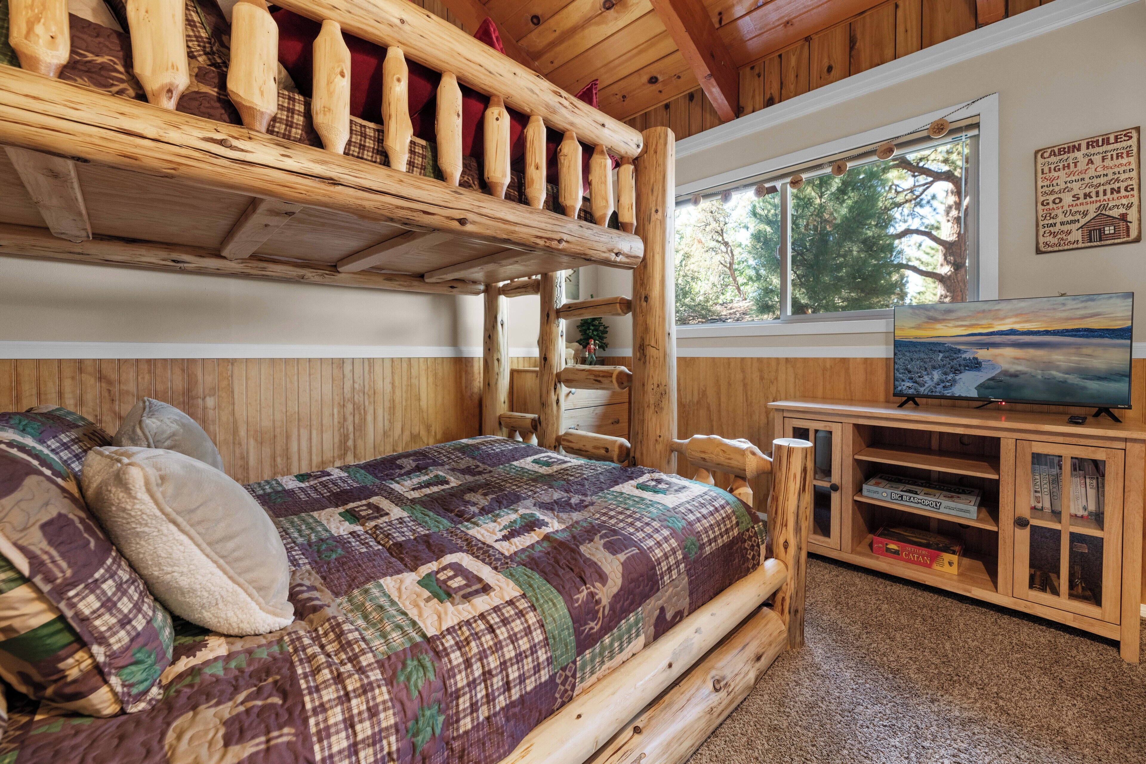 A cabin-style room with a bunk bed, a quilted bedspread, a television, and a wooden cabinet with books and games.
