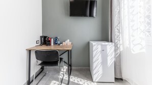 Une petite pièce comprenant un bureau, une chaise, un téléviseur à écran plat fixé au mur, un réfrigérateur et une fenêtre ornée de rideaux légers.