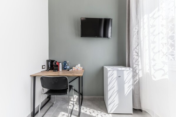 Una habitación pequeña con escritorio, silla, televisor de pantalla plana montado en la pared, refrigerador y ventana con cortinas translúcidas.