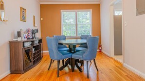 A dining area with a round table and four blue upholstered chairs, a wooden cabinet with kitchen items, and a large window with blinds. - MidTown Flats Suite C2 (Columbus)