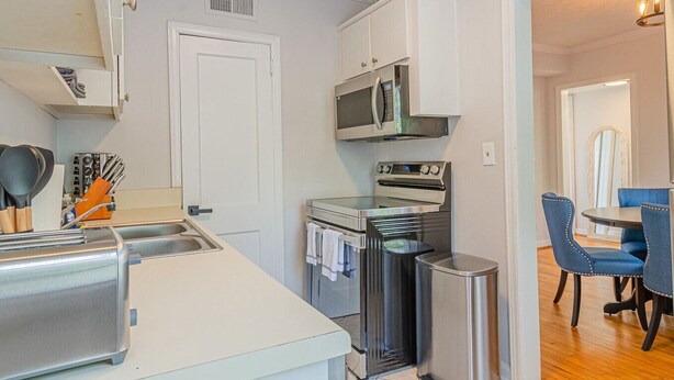 A compact kitchen with a microwave, oven, and sink. There is a dining area with blue chairs. - MidTown Flats Suite C2 (Columbus)
