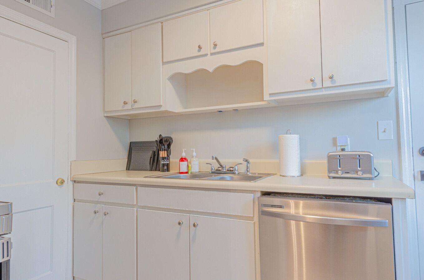 A kitchen with white cabinets, a stainless steel dishwasher, a sink with a faucet, a toaster, and a paper towel holder.
