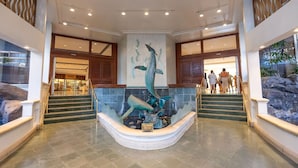 A lobby with a central aquarium featuring a large dolphin sculpture, a staircase with a railing, and a view of a shopping area in the background. - Steps to Beach  Resort Amenities  Kitchen (Honolulu)