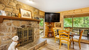 A cozy living room with a stone fireplace, wooden furniture, and a large window offering a view of the outdoors.