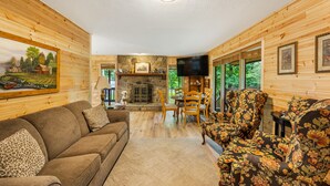 A cozy cabin interior with a stone fireplace, wooden walls, a sofa, floral armchairs, and a television.