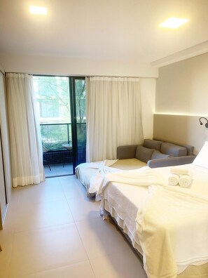 A hotel room with a bed, a sofa, a large window with curtains, and a view of greenery outside. - Beach Haus by Day by Day (Joao Pessoa)