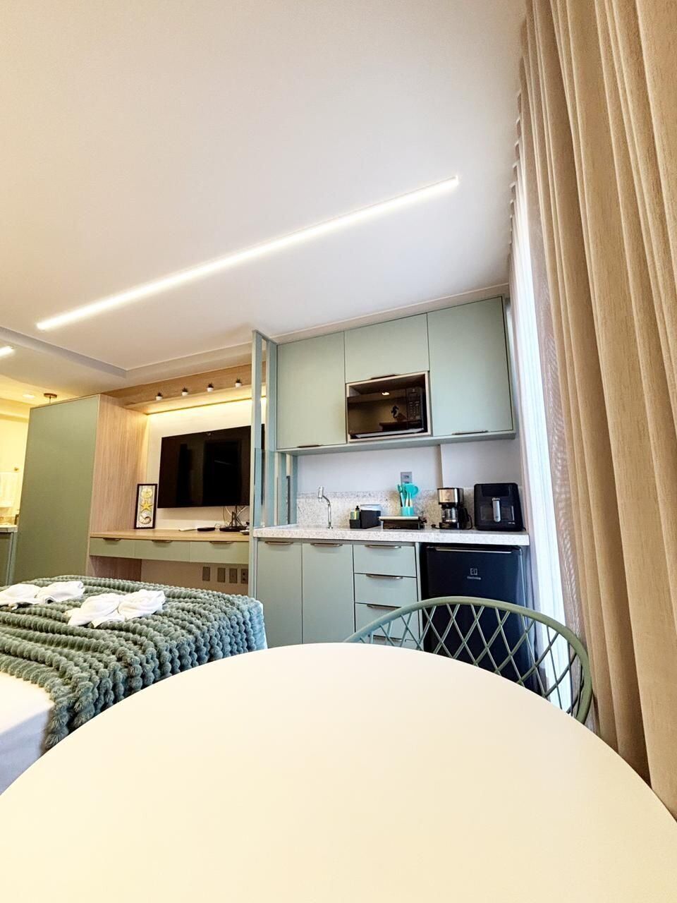 A compact hotel room with a kitchenette, a bed with a green blanket, and a window with curtains.