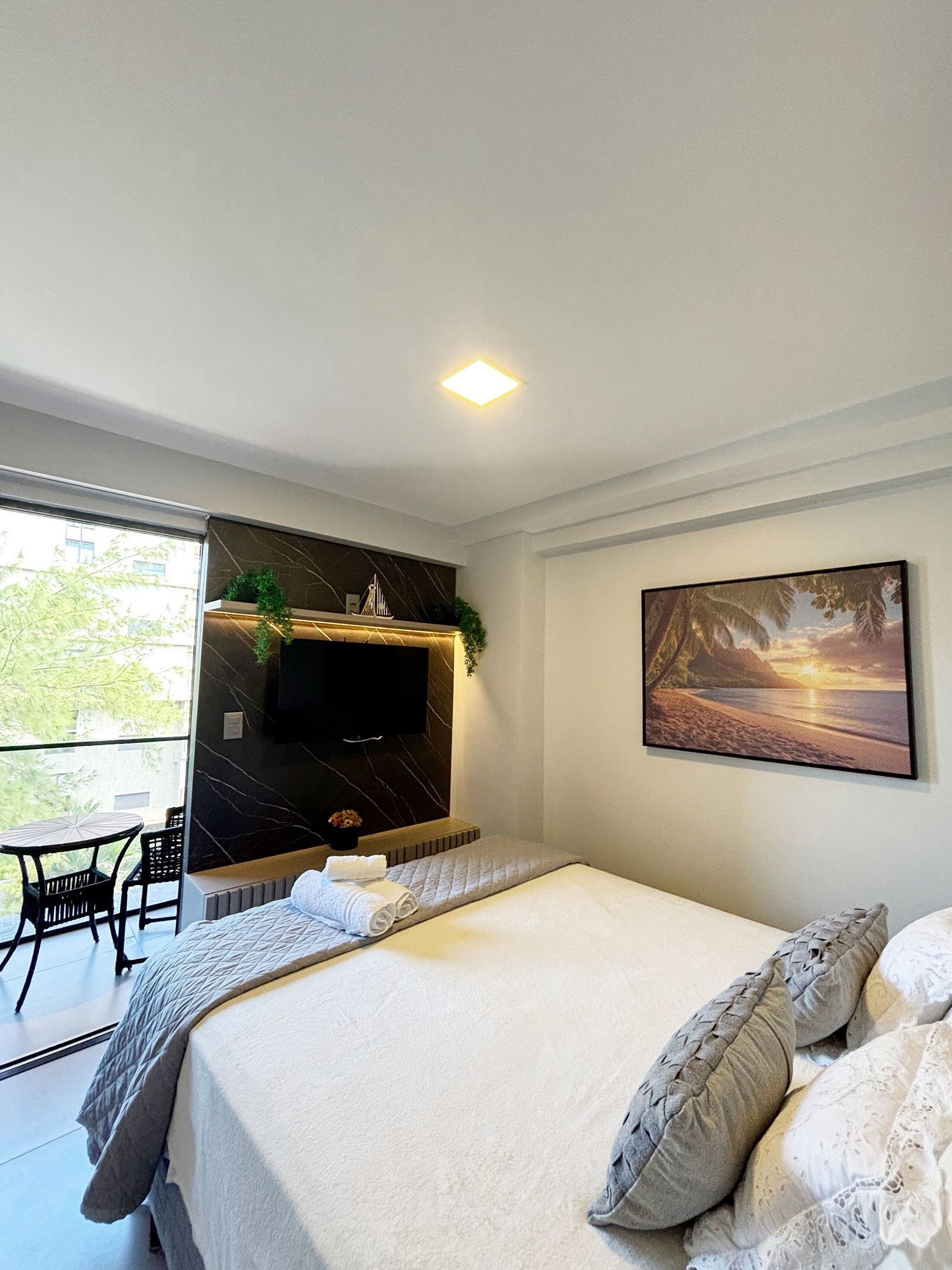 A modern bedroom with a bed, a TV mounted on a dark wall, a painting of a beach scene, and a balcony with a table and chairs.