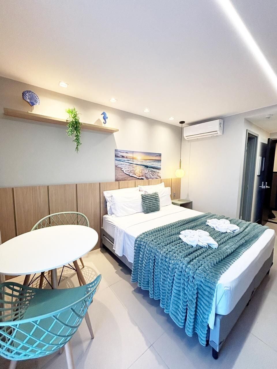 A hotel room with a bed, bedside table, chair, and a wall-mounted shelf with a plant.